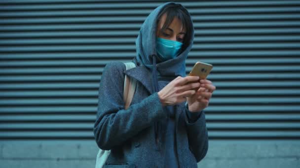 Mujer irreconocible en la capucha y la máscara facial contra el coronavirus de pie al aire libre utilizando el teléfono inteligente . — Vídeo de stock