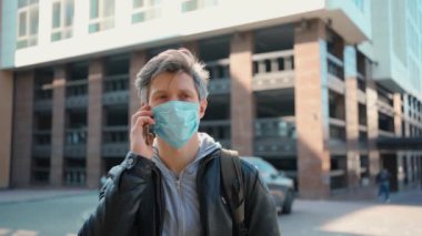 Koruyucu tıbbi maskeli gümüş saçlı adam dışarıda akıllı telefon kullanıyor..