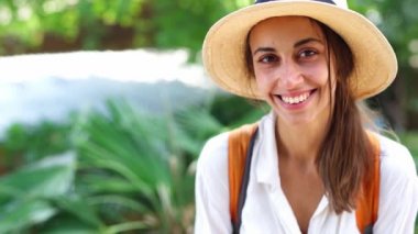 Hasır şapkalı, beyaz gömlekli güzel turist kadın kameraya bakıp gülümsüyor.