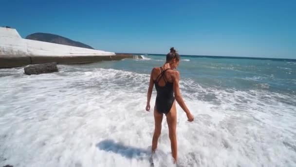 Alegre joven en traje de baño negro y gafas de sol de pie en el agua en la orilla del mar — Vídeos de Stock
