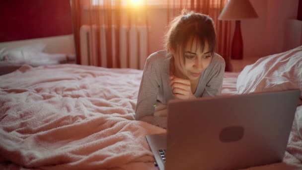 Junge Frau liegt zu Hause im Bett und schaut Laptop, surft im Internet. — Stockvideo