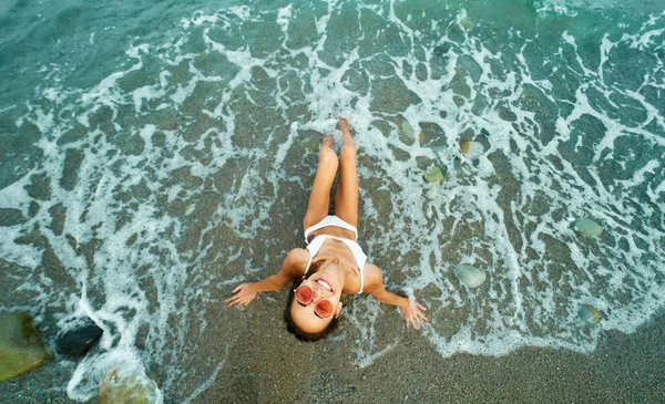 Vista superior chica divertida en gafas rojas se sienta en la playa con olas de mar alrededor . — Foto de Stock