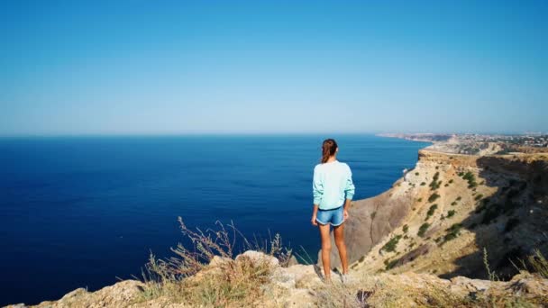 Vista trasera de la joven de pie en el borde del acantilado con hermosa vista al mar — Vídeos de Stock