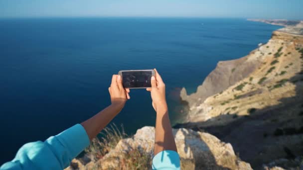 女性は携帯電話を手に取り美しい海の景色と崖の端で写真を撮る — ストック動画