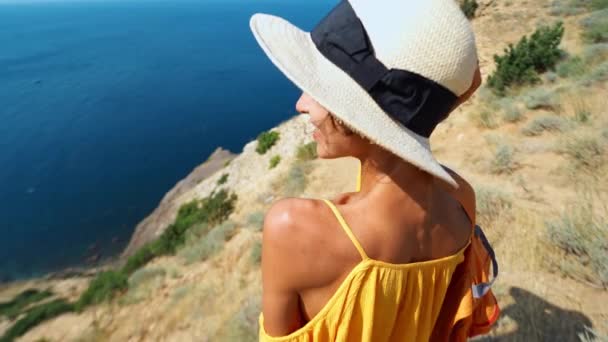 Kobieta w jasnożółtej sukience i kapeluszu z plecakiem stojącym na krawędzi klifu przed niesamowitym krajobrazem morskim — Wideo stockowe