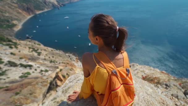 Sorridente ragazza in abito giallo e scarpe da ginnastica bianche seduta sul bordo della scogliera sopra la costa — Video Stock