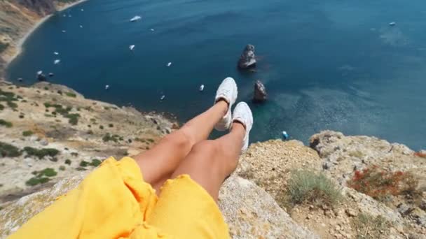 Camera zooming out first person view of taned woman legs in yellow dress sitting on on cliff edge with amazing nature view of seascape. — Stock Video