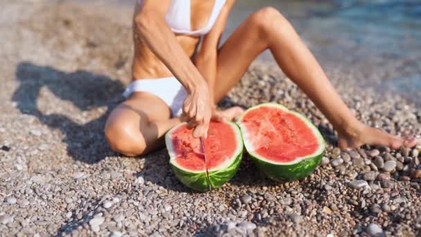 Fitbody woman in bikini sitting on pebble sea beach and cutting huge ripe watermelon — Stock Video