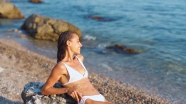Beyaz bikinili bronz tenli bir kadın, Pebble Beach 'te dinleniyor.