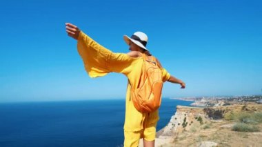 Sarı elbiseli ve şapkalı kadın deniz kıyısında uçurumun kenarına yürüyor.