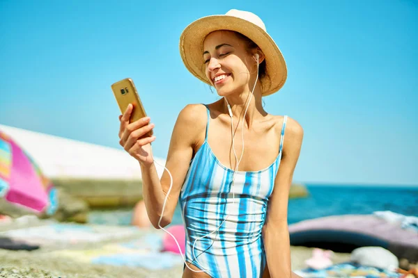 Lykkelig dame i stråhatt og badedrakt på stranden som kommuniserer på smarttelefon, nettbasert mobiltelefon. – stockfoto
