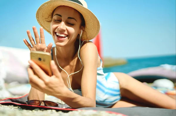 Ung dame med hatt ler og snakker i videomelding på smarttelefon, chatter med venner på stranden i sommerferien. – stockfoto