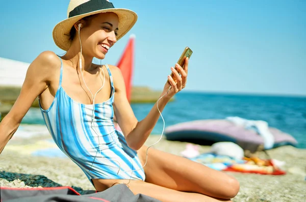 Радісна усміхнена дівчина в капелюсі лежить на пляжі на літніх канікулах, тримаючи смартфон — стокове фото