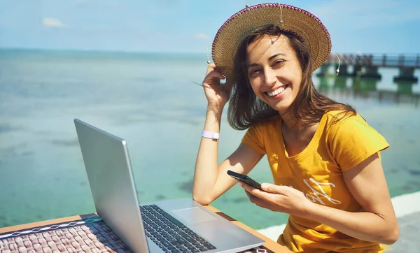 Muotokuva onnellinen freelancer nainen hattu hymyillen kameralle, tilalla älypuhelin, työskentelee kannettavan tietokoneen meren rannalla — kuvapankkivalokuva