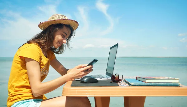 Frittgående kvinne med hatt som arbeider til sjøs ved hjelp av smarttelefon og internett. – stockfoto