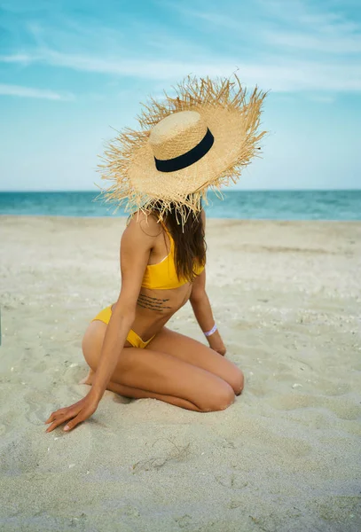 Pystysuora laukaus seksikäs bikinit ruskettunut elin nainen yllään iso olki hattu istuu hiekkarannalla sininen meri. — kuvapankkivalokuva