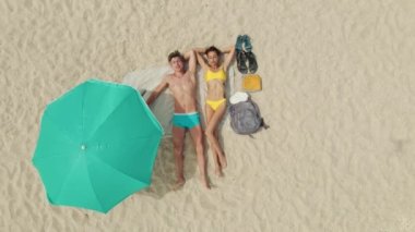 Turkuaz güneş şemsiyesinin altında, boş bir kumsalda beyaz kumların üzerinde uzanan çift.