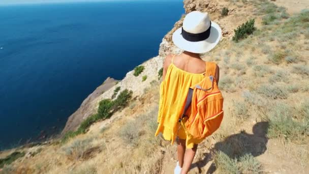 Caméra suivant femme en robe jaune et chapeau marchant au bord de la falaise sur le bord de la mer — Video