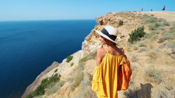Vrouw in helder gele jurk en hoed met rugzak staan op klif rand tegen verbazingwekkende zeegezicht — Stockvideo