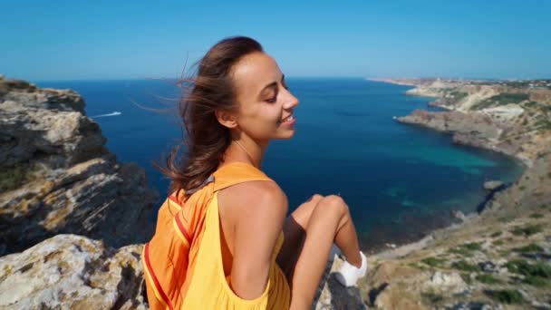 年轻纤细的女旅行者坐在悬崖顶上，面对着迷人的海景 — 图库视频影像