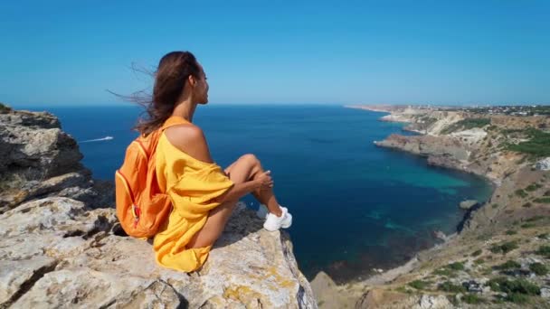Жінка сидить на вершині скелі, яскраво-жовта сукня і волосся, що дме на вітрі . — стокове відео