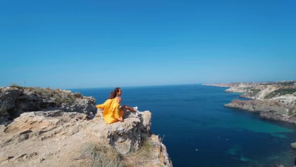 Kvinna sitter på klippan topp, ljus gul klänning och hår blåser i vinden. — Stockvideo