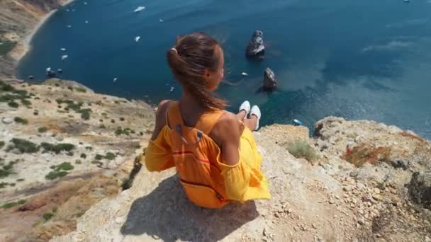 Usmívající se dívka ve žlutých šatech a bílé tenisky sedí na okraji útesu přes pobřeží — Stock video