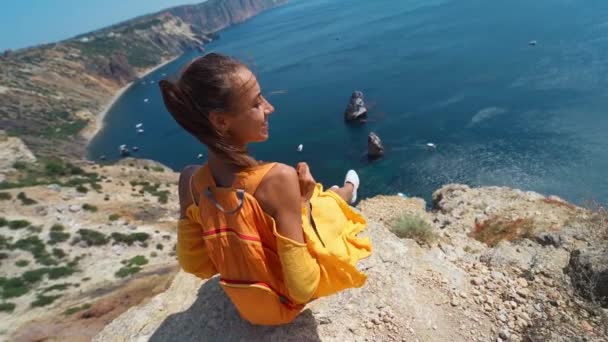Sorridente ragazza in abito giallo e scarpe da ginnastica bianche seduta sul bordo della scogliera sopra la costa — Video Stock