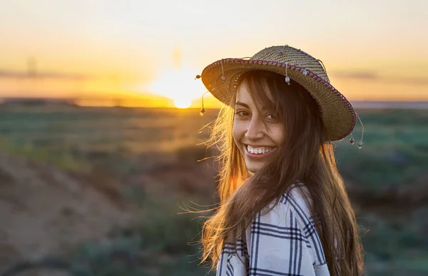 Utendørs portrett av en ung kvinne med hatt som smiler til kameraet i solstråler ved solnedgang – stockfoto