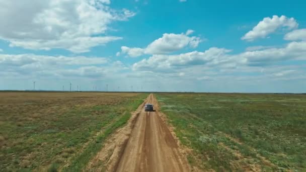Безпілотник після автомобіля, що рухається сільською дорогою через зелені поля на фоні вітрових турбін — стокове відео