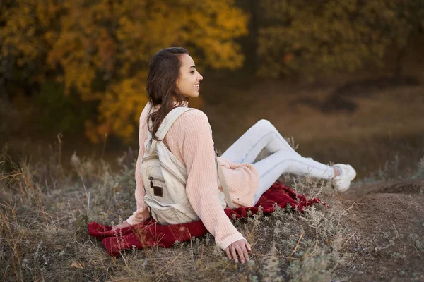 Kvinne i varm genser med ryggsekk sitter på teppe i høstparken med høst fargerik bakgrunn. – stockfoto