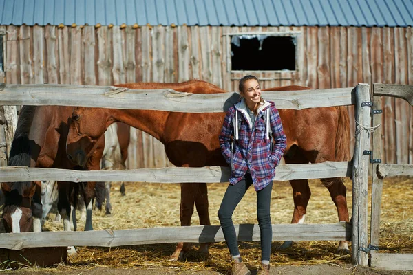 American female breeder standing by stable with horses on countryside farm or ranch. — Stock Photo, Image