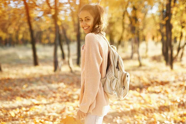 Chica bonita caminando en el parque de otoño con hojas caídas en las manos, follaje colorido y luces solares en el fondo. — Foto de Stock