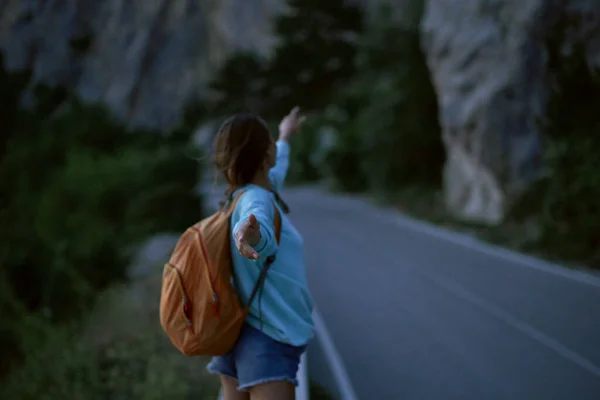Chica vista trasera con la mochila se para en la carretera con las montañas en el fondo por la noche — Foto de Stock