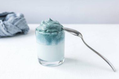 Mavi Matcha Dalgona, moda yumuşacık kremalı çırpılmış çay. Mavi kibrit çay, şeker ve sütten yapılmış bir içecek. Cam beyaz masa, gri peçete ve kaşık. Yumuşak odaklanma. Kapat.. 