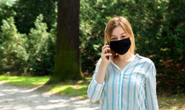 Siyah koruyucu maskeli beyaz bir kadın. Parkta telefonla konuşuyor.