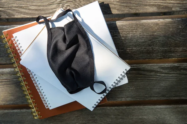 Máscara protectora negro en los cuadernos en un banco del parque. — Foto de Stock