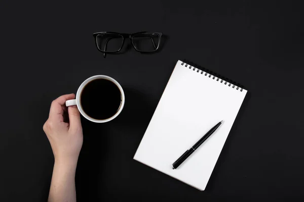 Mano sostiene una taza de café en una mesa negra de trabajo al lado del bloc de notas — Foto de Stock