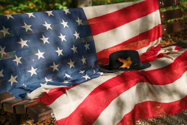 Sombrero negro con una hoja de arce amarillo en un banco con una bandera americana —  Fotos de Stock