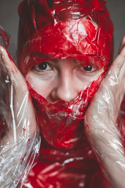 Screaming human face pressing through plastic stretch membrane painted red color as horror background. close up