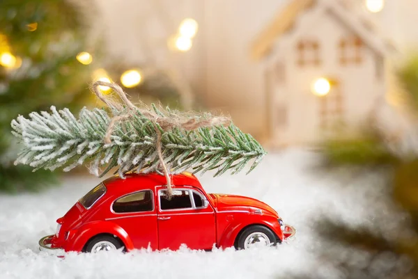 Christmas red toy car delivering fir tree with house and garland bokeh lights on the background. New year greeting card. Winter holiday