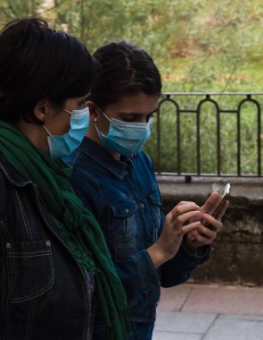 Kot ceketli ve maskeli bir kız, 19. periyotta şehirde yürürken annesine cep telefonunu ve uygulamaları gösteriyor.
