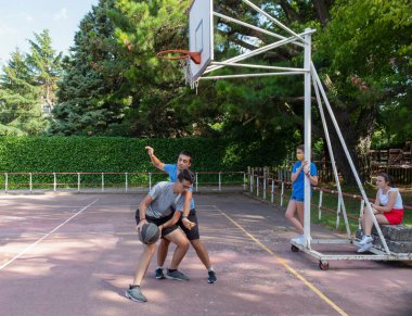 Açık hava basketbol sahasında basketbol oynayan iki oğlan ve kovid19 zamanında onları maske takarak izleyen iki kız.