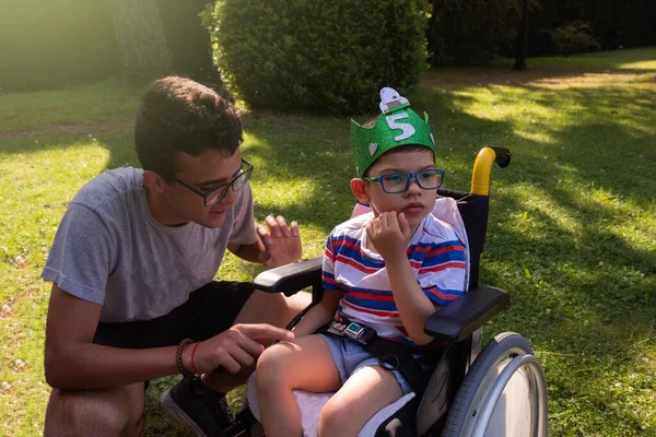 Tekerlekli sandalyedeki çocuk Pitthopkins sendromu ile el yapımı bir Eva kauçuk çelengi ve ona bakan bir ergenle bahçede doğum gününü kutluyor.