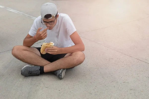 夏の日に太陽から彼を守るために帽子でアスファルトの床に座ってジャガイモとパンとトルティーヤサンドイッチを食べる若い男 — ストック写真