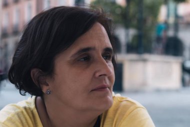 Düşünceli, yorgun, orta yaşlı, sarı tişörtlü bir kadın yaz gününde aşağıya bakıyor.