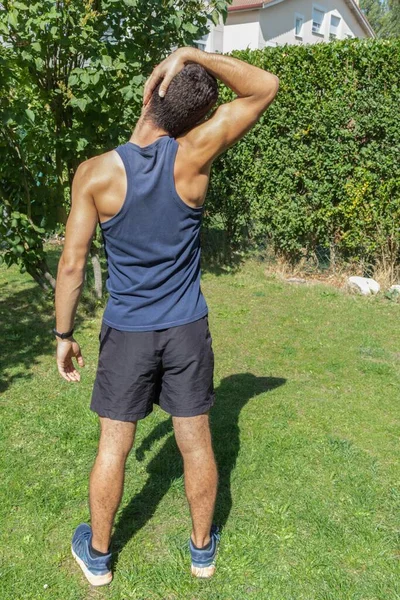 Spor kıyafetli genç bir çocuk güneşli bir günde evin bahçesinde spor yapmak için boynunu esnetiyor.