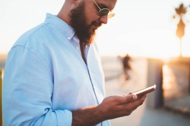 Erkek hipster haber online holding smartphone posta denetimi metin mesajı ve şehir üzerinde cep telefonu app duran üzerinde sokak günbatımında gönderme sokak, şık yakışıklı delikanlı okuma görünümünü kırpılmış