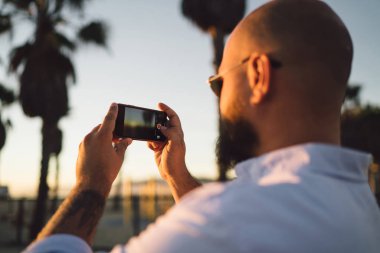 Dikiz genç erkek tutan eller smartphone ile modern fotoğraf makinesi için güzel gün batımı, fotoğraflarını alarak kırpılmış hipster adam cep telefonu üzerinde akşam manzara çekim görünümünü yakın