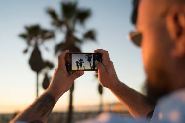 Erkek turist çekim güzel sahne günbatımı kullanarak video görünümü kadar yakın smartphone kamera, fotoğraf ve egzotik palmiye ağaçlarının akşamları çekmek için modern telefon hipster adam görünümünü kırpılmış